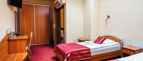 A hotel room with a bed, bedside tables, a desk with a chair, a TV, and an air conditioning unit.