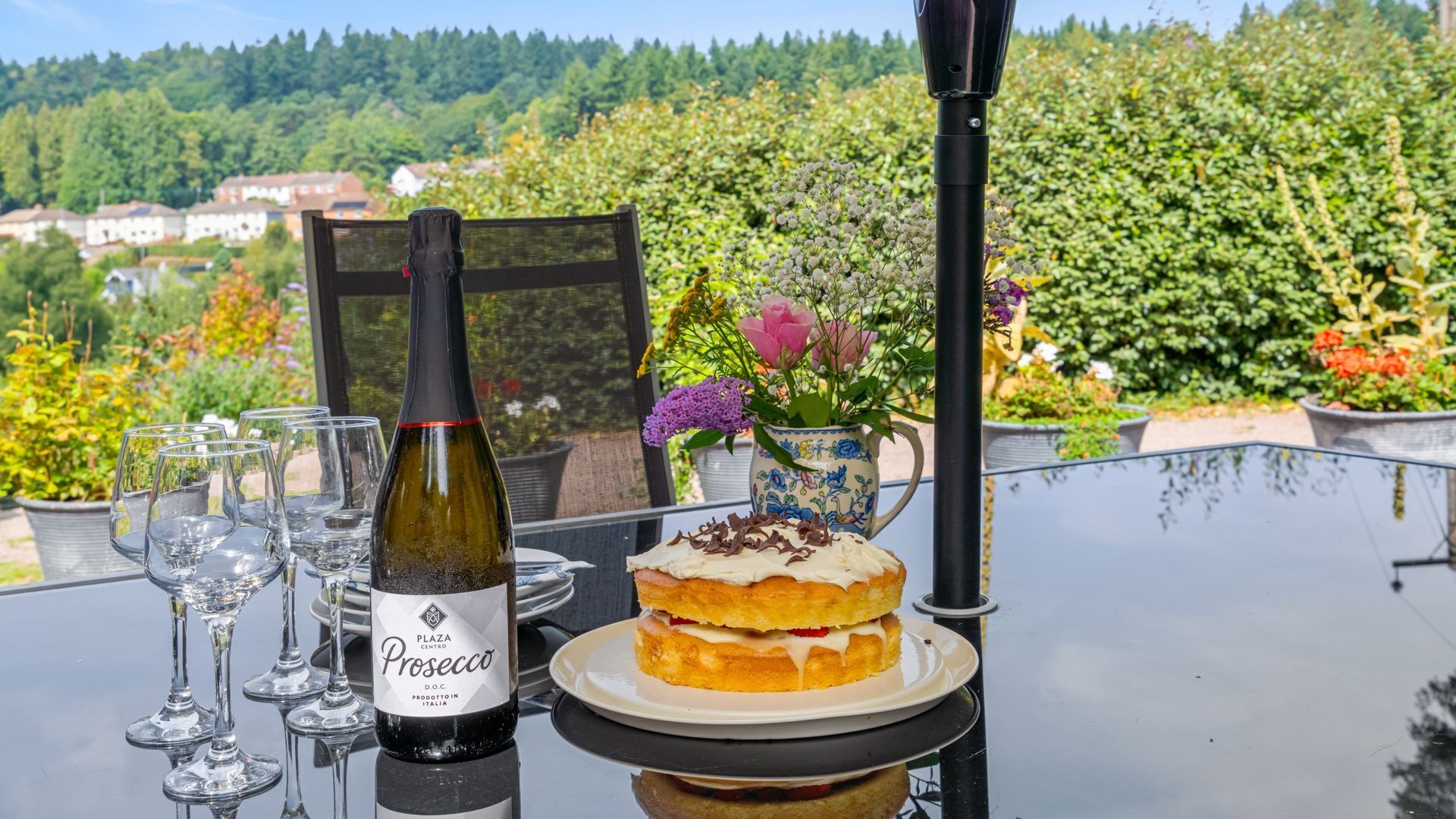 A table with a bottle of Prosecco, glasses, and a dessert on a terrace with a scenic view.