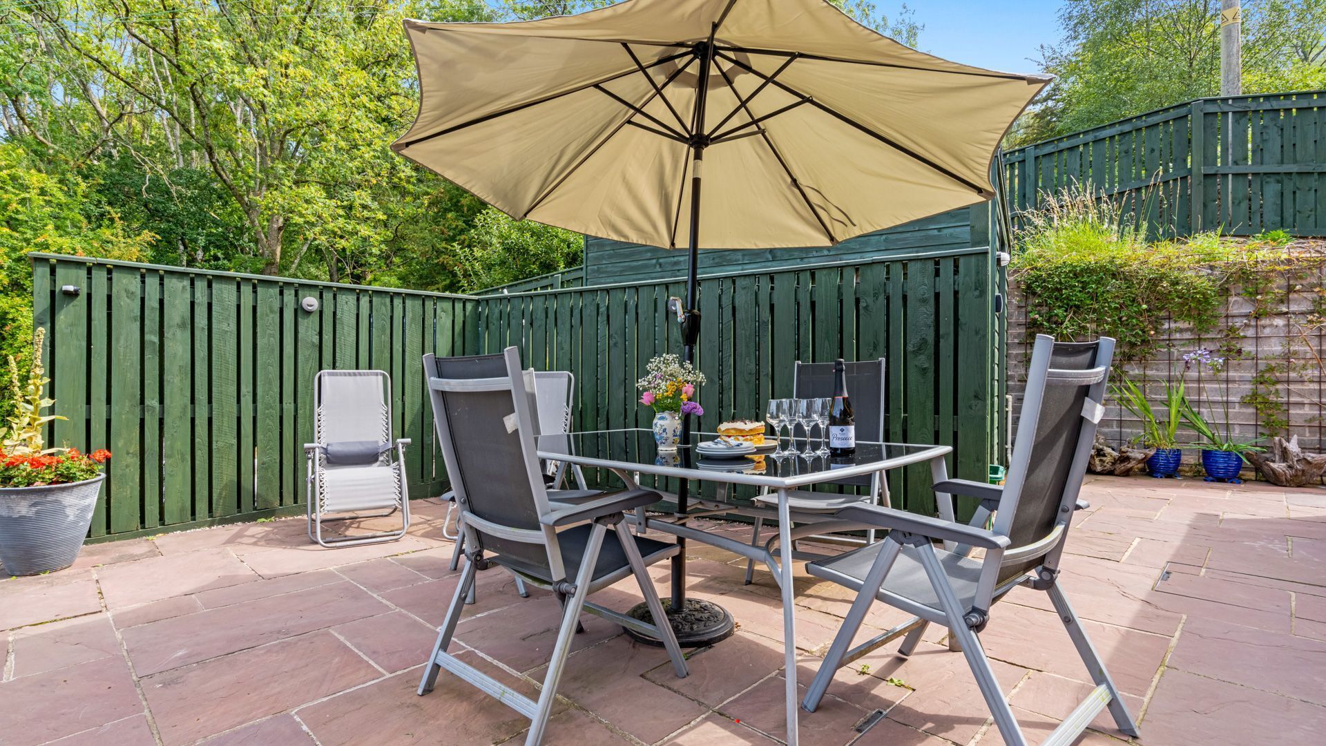 Une terrasse extérieure avec une table et des chaises, un grand parasol et une clôture verte.
