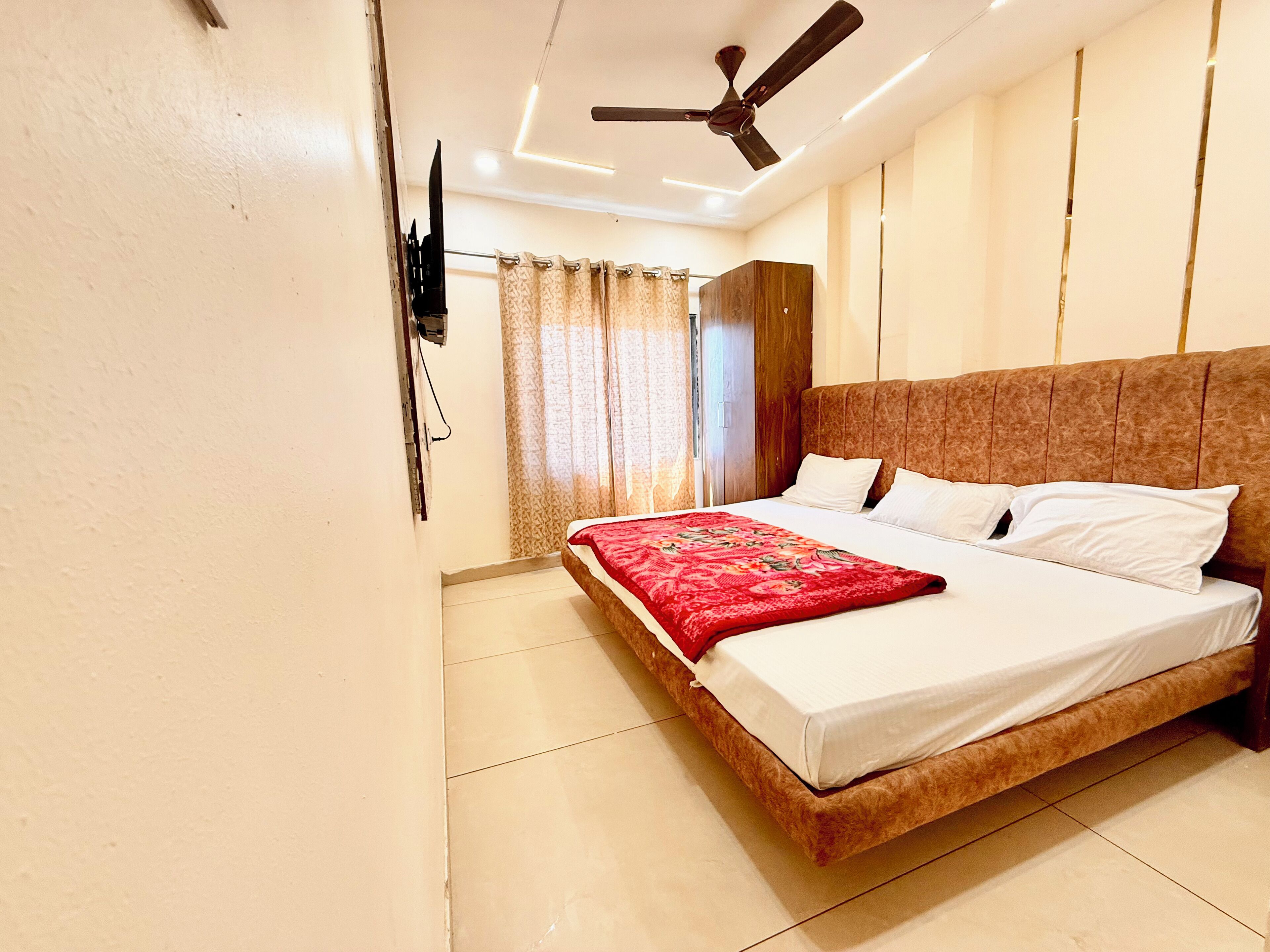A bedroom with a bed, a ceiling fan, a TV mounted on the wall, and a window with curtains.