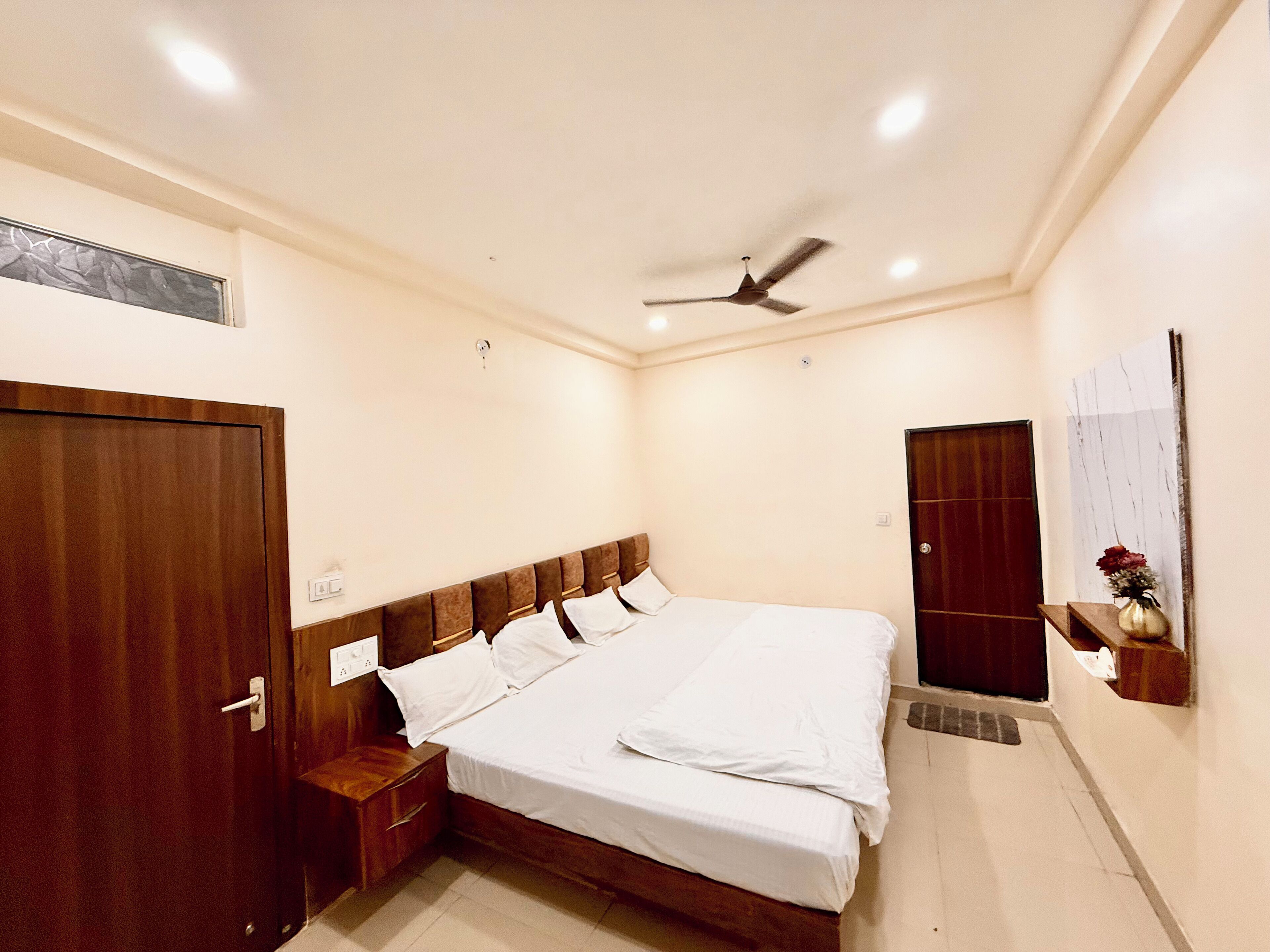 A bedroom with a large bed, wooden headboard, a ceiling fan, a wooden door, and a wall-mounted shelf with a vase.