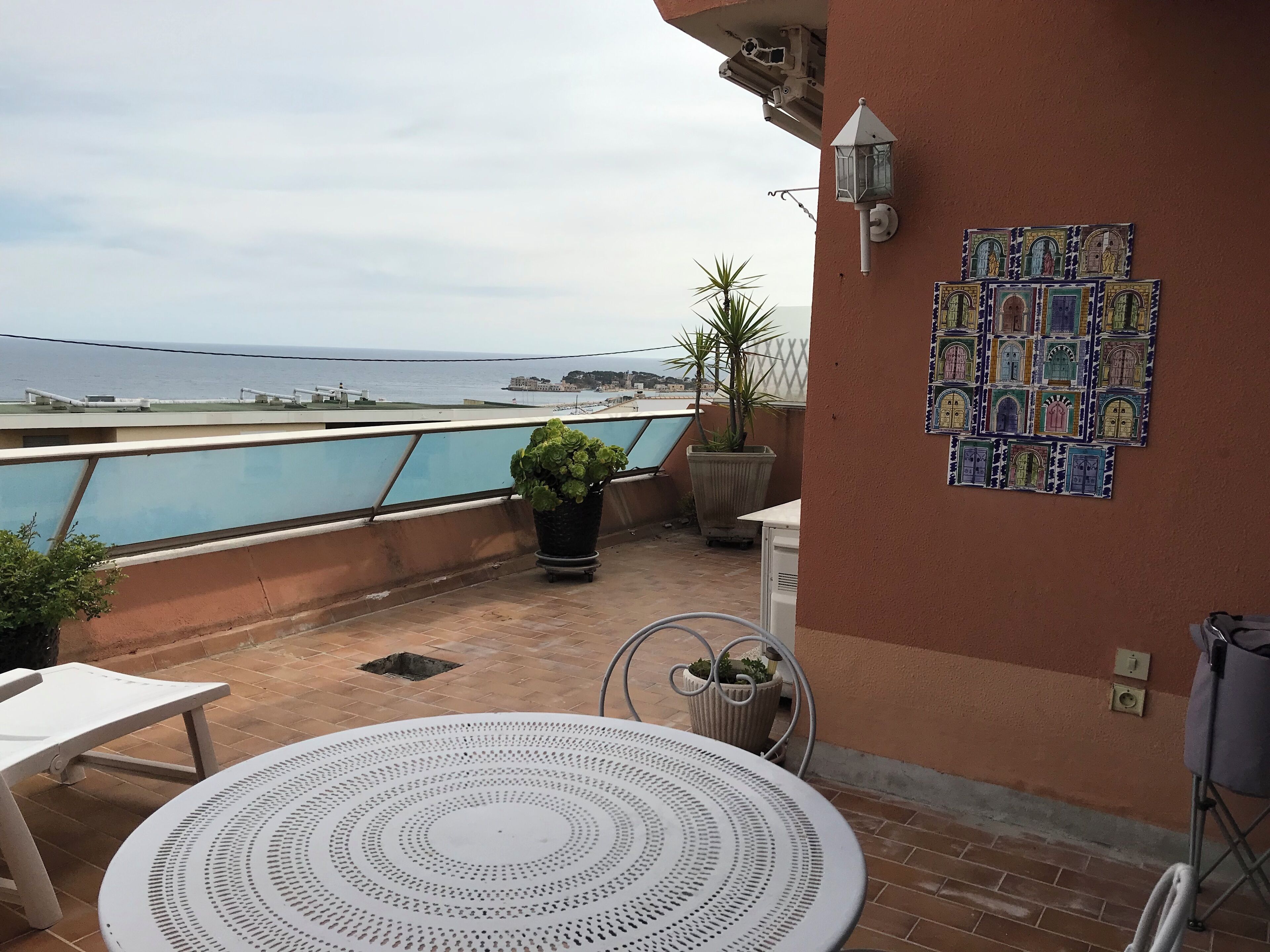 A rooftop terrace with a view of the sea, a potted plant, a table, and a decorative wall piece.