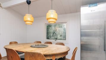 A dining area with a round wooden table, six wooden chairs, two hanging pendant lights, a stainless steel refrigerator, and a framed painting on the wall.