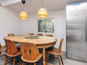A dining area with a round wooden table, six wooden chairs, two hanging pendant lights, a stainless steel refrigerator, and a framed painting on the wall. - 4 Star Holiday Home in Hornbaek (Hornbaek)