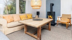 A cozy living room with a sofa, a wooden table, a chair, a fireplace, and a large window. - 4 Star Holiday Home in Hornbaek (Hornbaek)