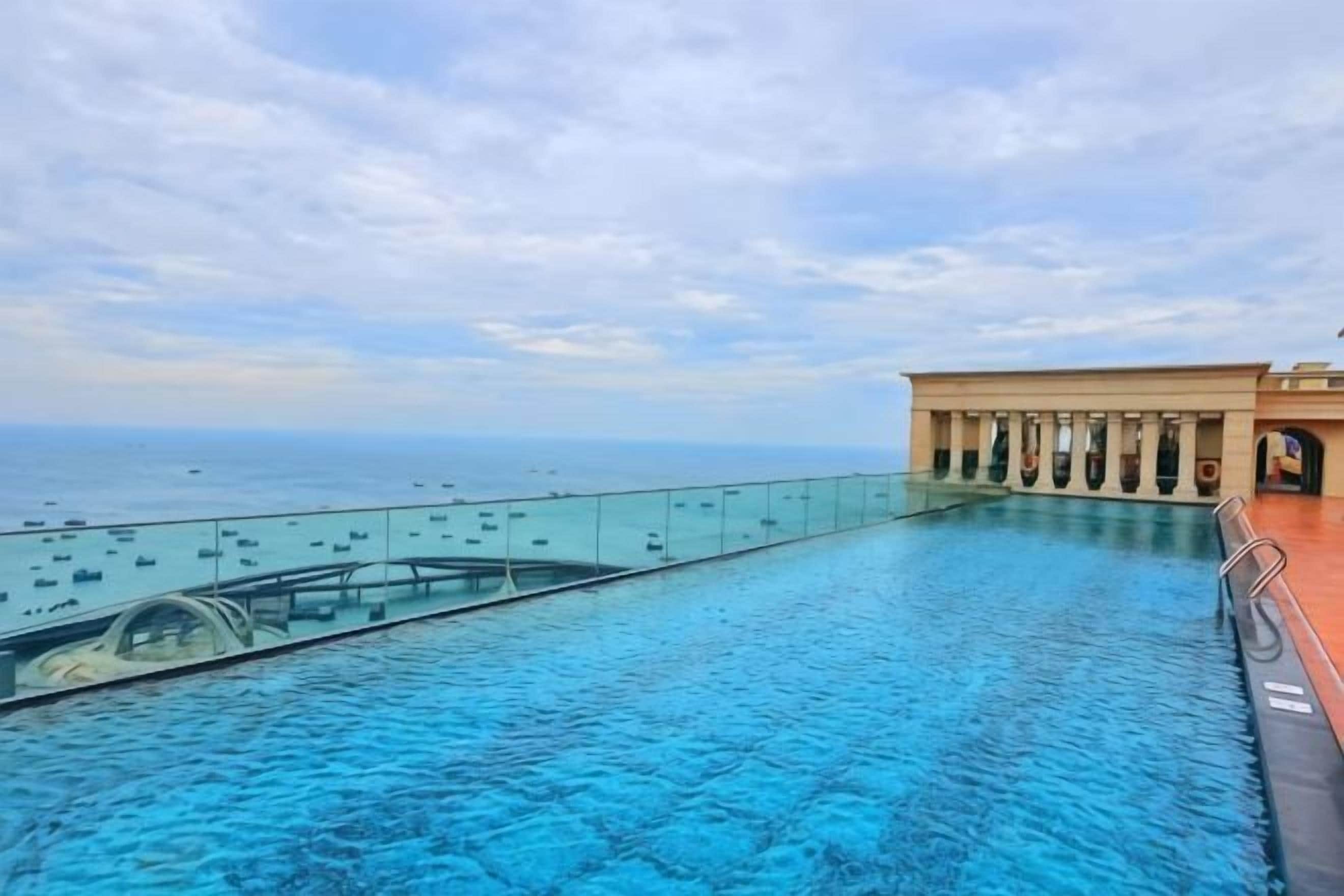 Une piscine avec une paroi en verre donnant sur la mer, un bâtiment orné de colonnes et un ciel dégagé.