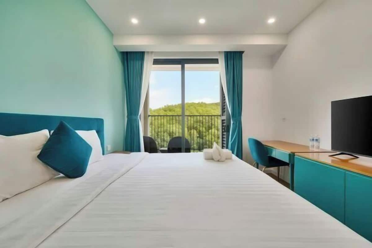 Une chambre d’hôtel équipée d’un lit, d’un bureau, d’une chaise, d’une télévision et d’un balcon donnant sur un paysage verdoyant.