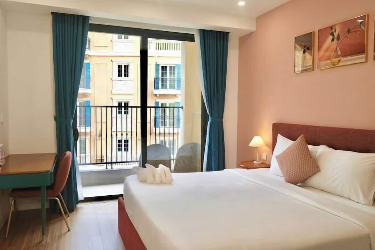 Une chambre d’hôtel comprenant un lit, un bureau, une chaise, une table de chevet avec une lampe, et une vue sur les bâtiments depuis un balcon.