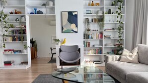 A modern living room with a glass coffee table, a grey armchair, and a grey sofa. There are white bookshelves filled with books and plants, and a painting on the wall. - Caves Beach, Sea Breeze - 5 min walk to the patrolled beach and caves (Caves Beach)