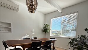A dining area with a wooden table and chairs, a chandelier, a large window with blinds, and a potted plant. - Caves Beach, Sea Breeze - 5 min walk to the patrolled beach and caves (Caves Beach)
