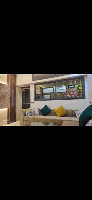 Een moderne woonkamer met een beige bank, groene en gele kussens, en een groot raam met uitzicht op groen.