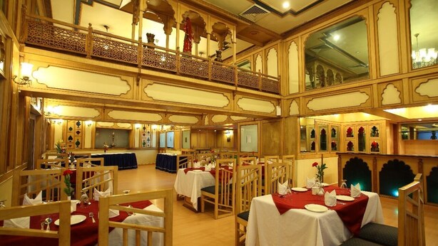 A dining area with multiple tables set for service, a balcony with ornate railings, and a high ceiling with decorative moldings. - Hotel Om Tower & Revolving Restaurant  (Jaipur)