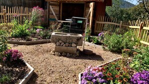 Une cabane en bois rustique, dotée d’un puits en pierre, entourée de parterres surélevés où poussent diverses plantes et fleurs.