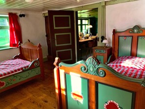 A room with two wooden beds, a wooden chair, a desk with a lamp, and a door leading to another room.