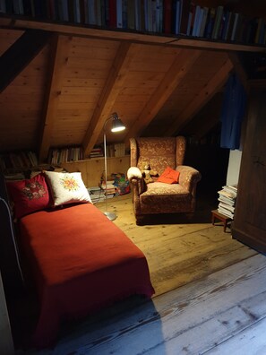 A cozy attic room with a bed, a chair, a lamp, and a bookshelf filled with books.