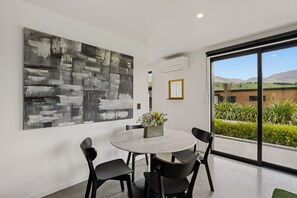 A modern dining area with a round table, four black chairs, a large abstract painting, a small vase with flowers, and a view of a building and hills through large glass doors. - Luxury Coronet Residence (Queenstown)