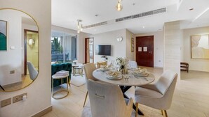 A modern dining area with a round table set for four, surrounded by upholstered chairs. A television is mounted on the wall, and there is a small round table with a vase of flowers. -  Durrani Homes - Azure 2BR with Burj Khalifa and Fountain View (Dubai)