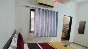 A hotel room with a bed, a window with curtains, an air conditioning unit, and a door leading to another room. - HOTEL ARADHANA (Veraval)