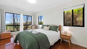 A bedroom with a large bed, a nightstand, a chair, and a view of trees through the windows. - Havenwood Chalet 2 Bedrooms (Ravensbourne)