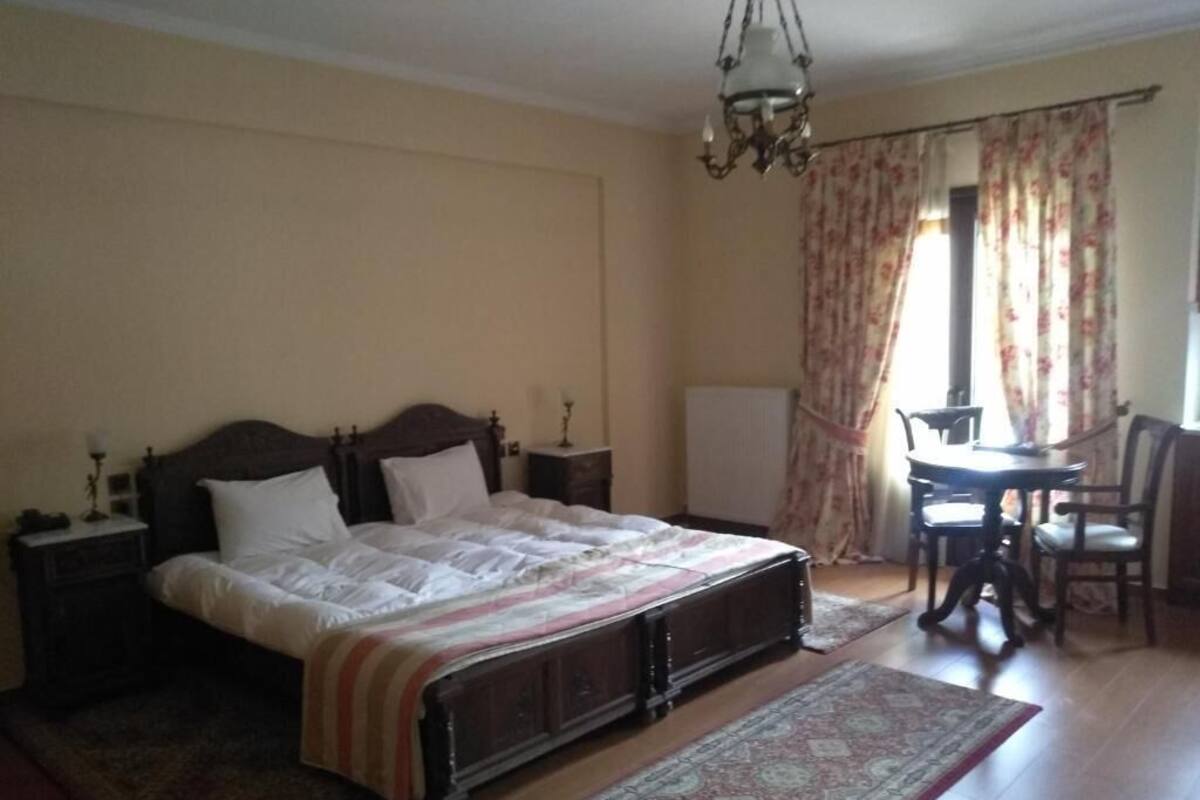 A bedroom with a large bed, a nightstand, a table with chairs, and a window with curtains.