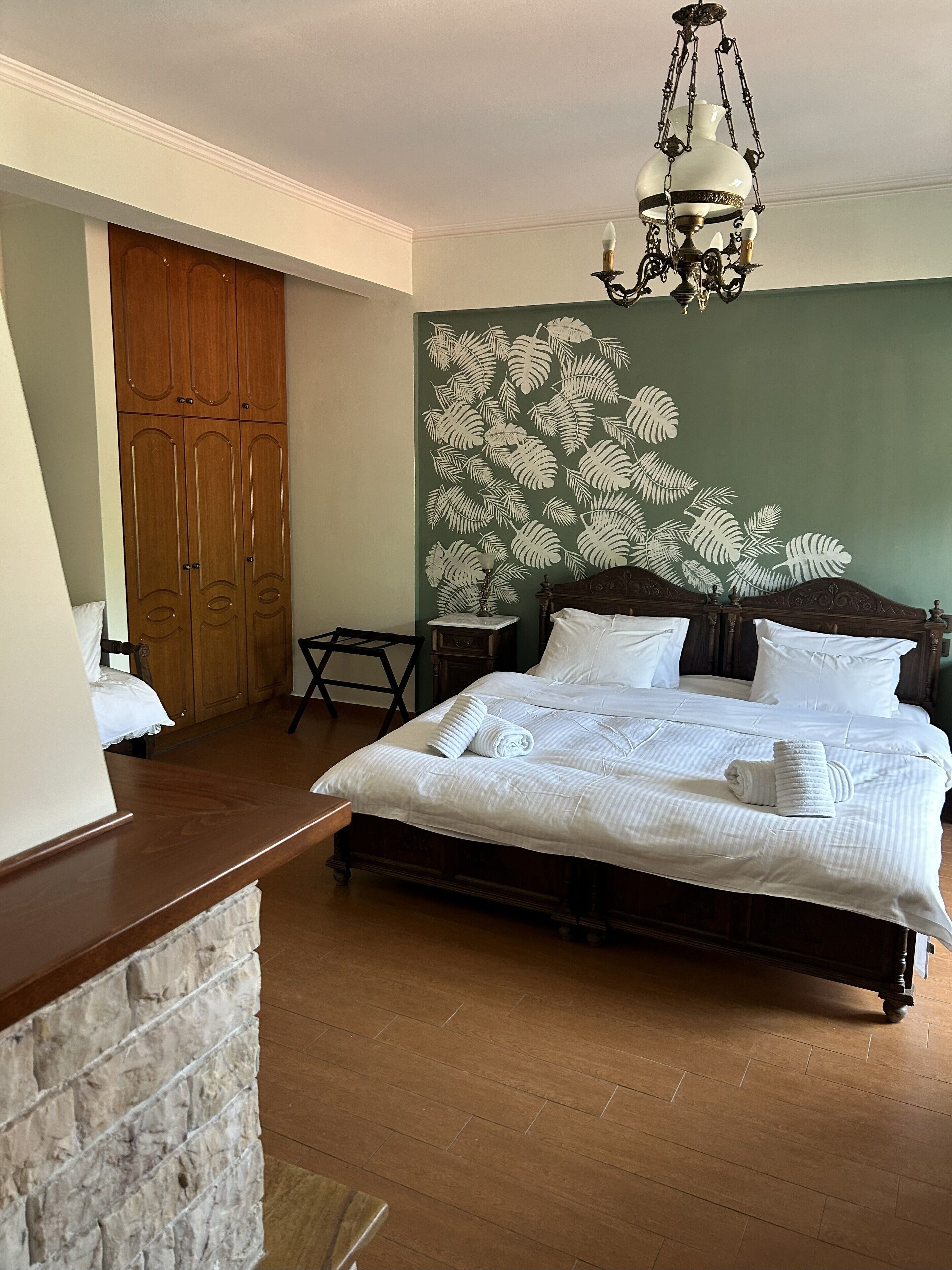 A neatly made bed with white linens and pillows, a wooden headboard, a nightstand with a lamp, and a large fern leaf wall mural.