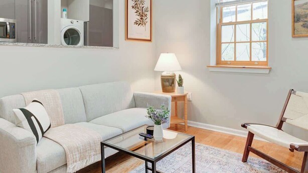 A living room with a sofa, a coffee table, a chair, a lamp, a window, and a kitchen area with appliances. - Cozy 1BD In The Heart Of Rittenhouse Square (Philadelphia)
