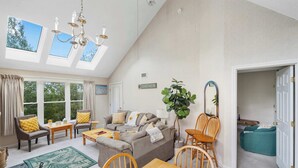 A spacious living area with a high ceiling, skylights, a chandelier, a sofa set, a dining table, and a view of the outdoors.