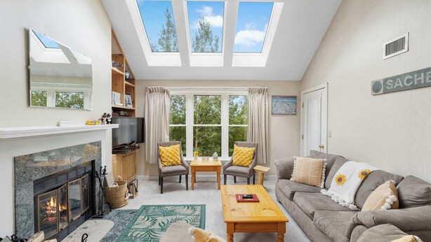 A living room with a fireplace, sofa, coffee table, and a television. There are skylights, a window, and a door with a sign.