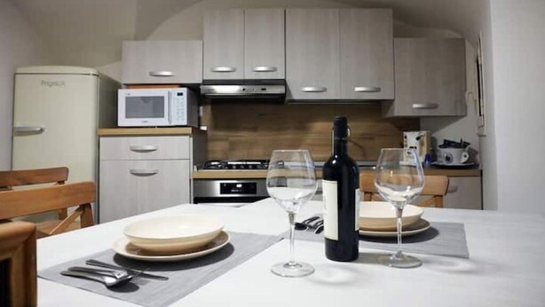 Un coin repas avec une table dressée pour deux personnes, une bouteille de vin et deux verres à vin. On aperçoit un four à micro-ondes et une cuisinière au fond.