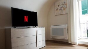 Un salon moderne avec un téléviseur à écran plat affichant le logo de Netflix, un meuble blanc avec des tiroirs, un radiateur et une fenêtre avec des rideaux.