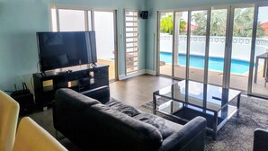 Una sala de estar con televisor de pantalla plana, un sofá de cuero negro y una mesa de centro de vidrio. Hay una puerta corrediza de vidrio que da a una zona de piscina.