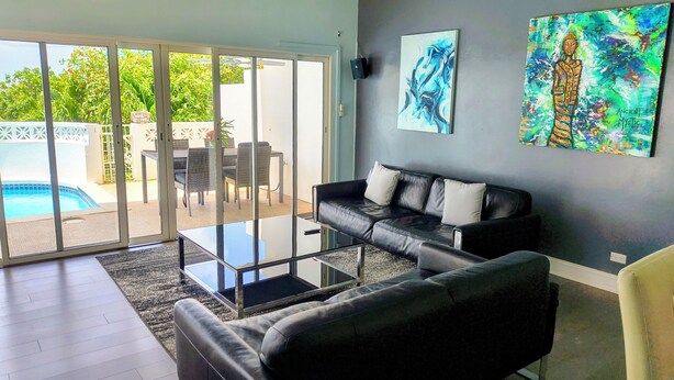 Una sala de estar moderna con un sofá de cuero negro, una mesa de centro de vidrio y una piscina visible a través de puertas de cristal.