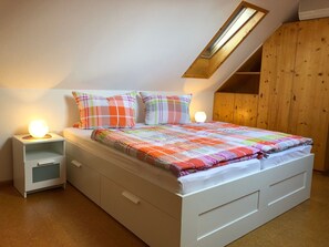 Une chambre à coucher avec un plafond en pente, un lit recouvert d’une courtepointe aux couleurs vives, deux lampes de chevet et une armoire en bois.