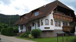 Une maison à deux étages avec un toit en pente, un balcon doté de balustrades en bois et un petit jardin agrémenté de diverses plantes et fleurs.