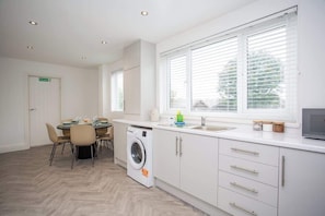 A modern kitchen with a washing machine, a dining area with chairs, and a large window with blinds.