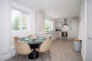 A modern kitchen with white cabinets, a dining area with a round table and chairs, and a view of the outdoors through large windows.