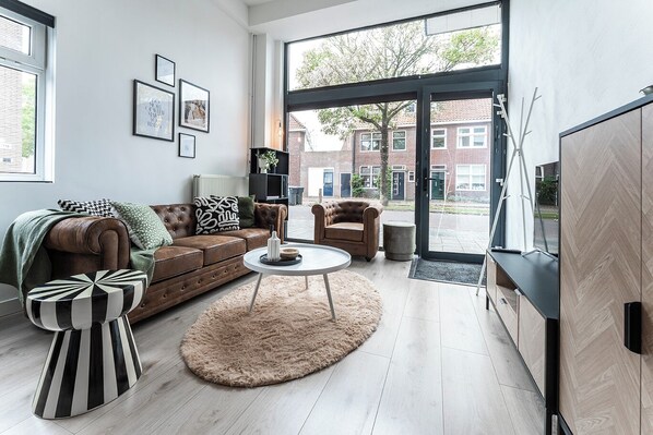 A modern living room with a brown leather sofa, a round coffee table, and a large window offering a view of a brick building. - Stylish and spacious open loft with double sleeping areas  in the heart of Tilburg. Modern, bright, and fully equipped with kitchen, fast WiFi, smart TV, and workspace — perfect for a city getaway or comfortable workation. (Tilburg)