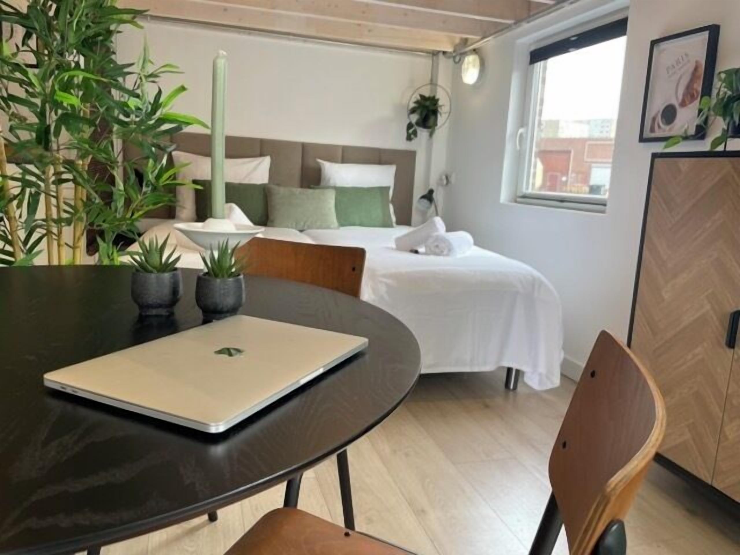 A modern hotel room with a bed, a desk with a laptop, a chair, and potted plants.