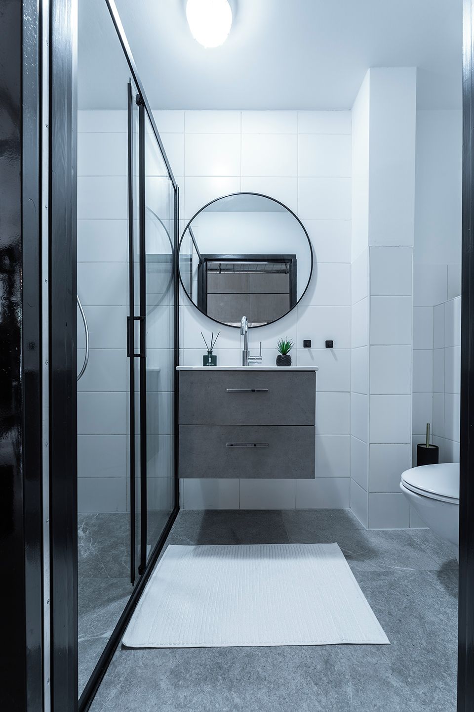 Un baño moderno con cabina de ducha de vidrio, un espejo redondo, un mueble de tocador gris y un inodoro blanco.