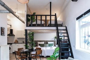 A compact studio apartment with a kitchenette, a dining area, a bed with a headboard, and a staircase to the upper bunk.