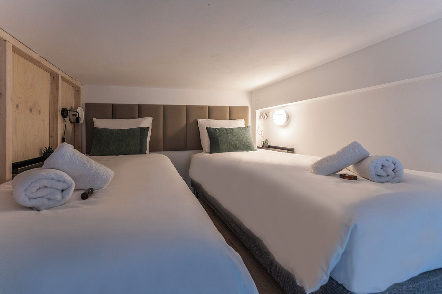 Habitación de hotel con dos camas, cada una con sábanas blancas y almohadas verdes, un armario de madera y una mesita de noche con lámpara.