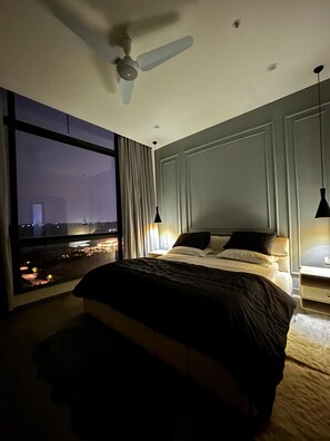 A hotel room with a large bed, a ceiling fan, a nightstand with a lamp, and a view of a cityscape through a large window. - Chic & Cozy 2BR Apt Premium Massage Chair Skypark1 Gulberg Greens Islamabad (Islamabad)