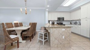 A kitchen with white cabinets, a microwave, stove, and a dining area with wicker chairs and a white table. - Lazy River | 15th Floor Views | Beachfront Condo (Orange Beach)