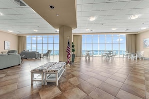 A spacious indoor area with a large window offering a view of the ocean, a flag, and a seating area with a bench and chairs. - Lazy River | 15th Floor Views | Beachfront Condo (Orange Beach)