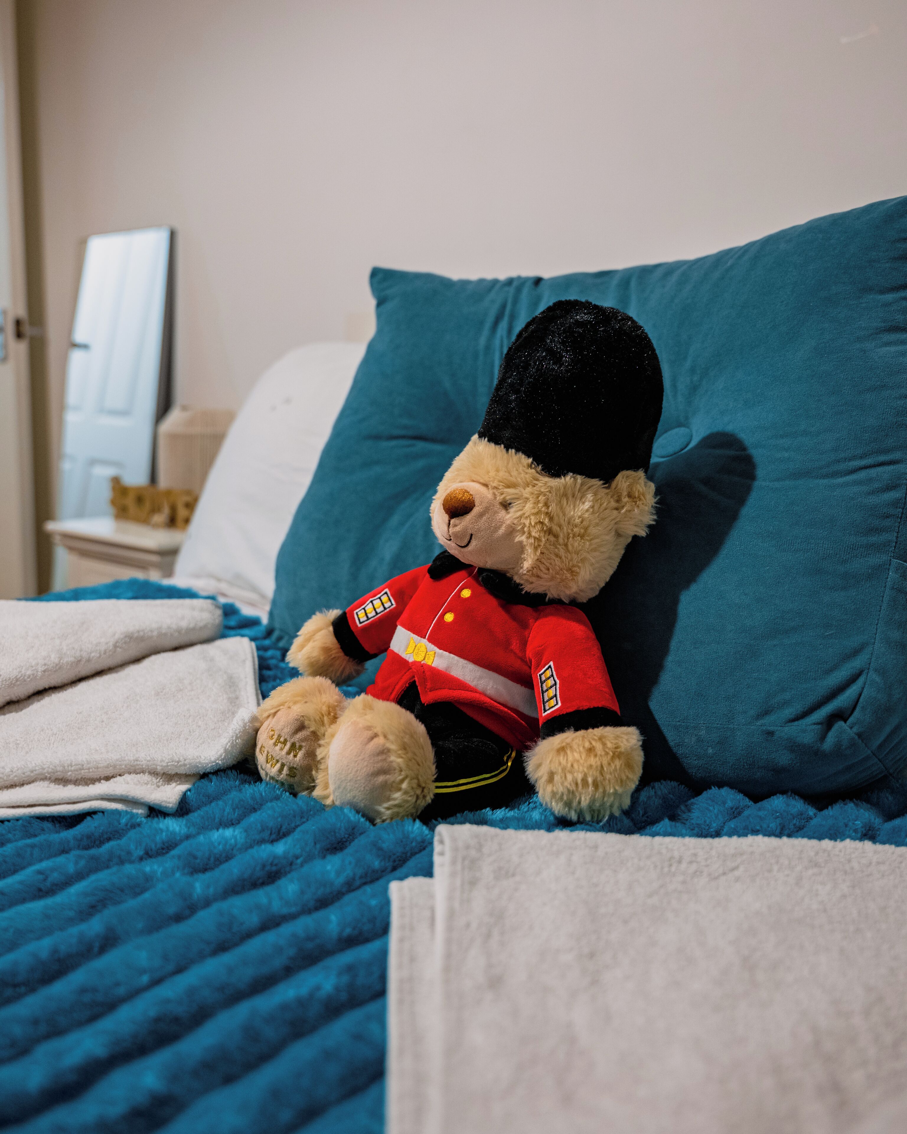 A teddy bear in a uniform sitting on a bed with a blue pillow and white sheets.
