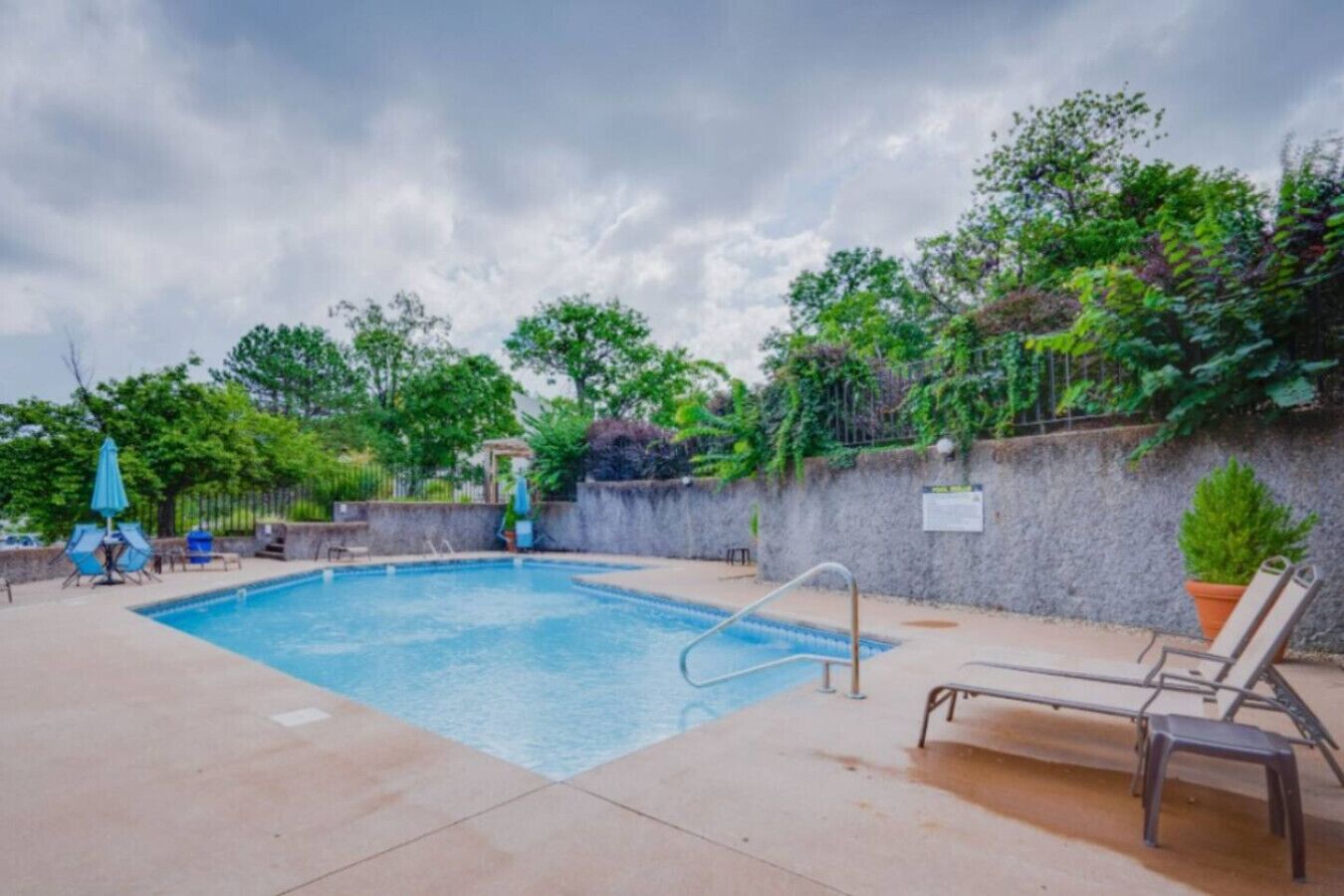 A rectangular swimming pool with a ladder, surrounded by a concrete deck. There are lounge chairs and a small table with umbrellas.