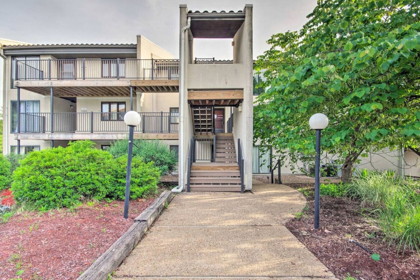 A multi-story residential building with a covered staircase, a balcony, and a garden area with shrubs and a tree.