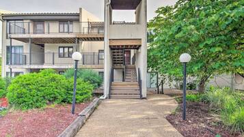 A multi-story residential building with a covered staircase, a balcony, and a garden area with shrubs and a tree.