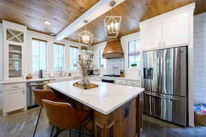 A modern kitchen with white cabinetry, a large island, stainless steel appliances, and wooden ceiling beams. - Sugarberry Farm | Farmhouse | hot tub | extra beds (Broken Bow)
