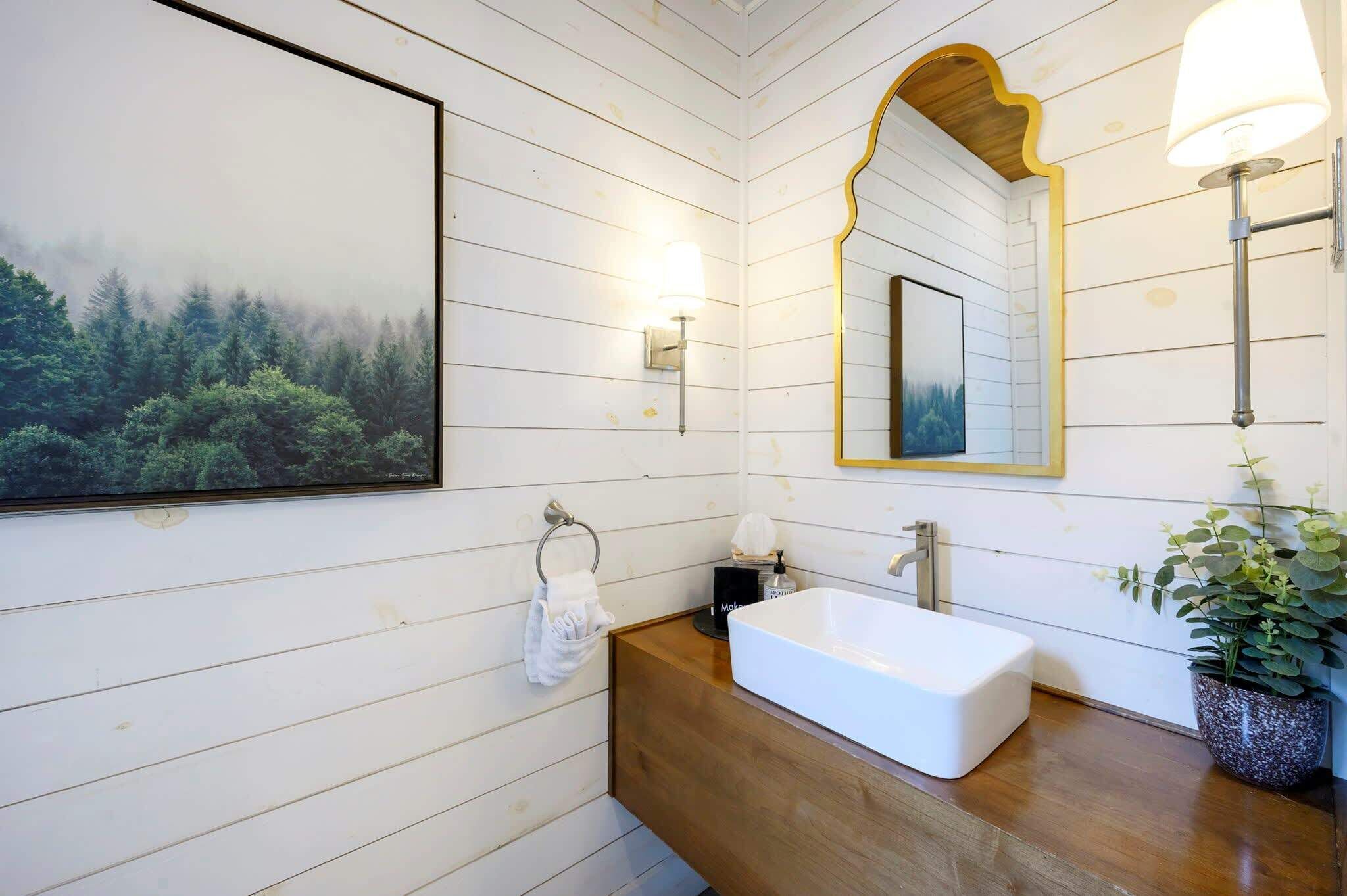 Une salle de bain avec un lavabo blanc, un meuble-lavabo en bois, un tableau encadré représentant une forêt, un miroir avec un cadre doré et une plante en pot.